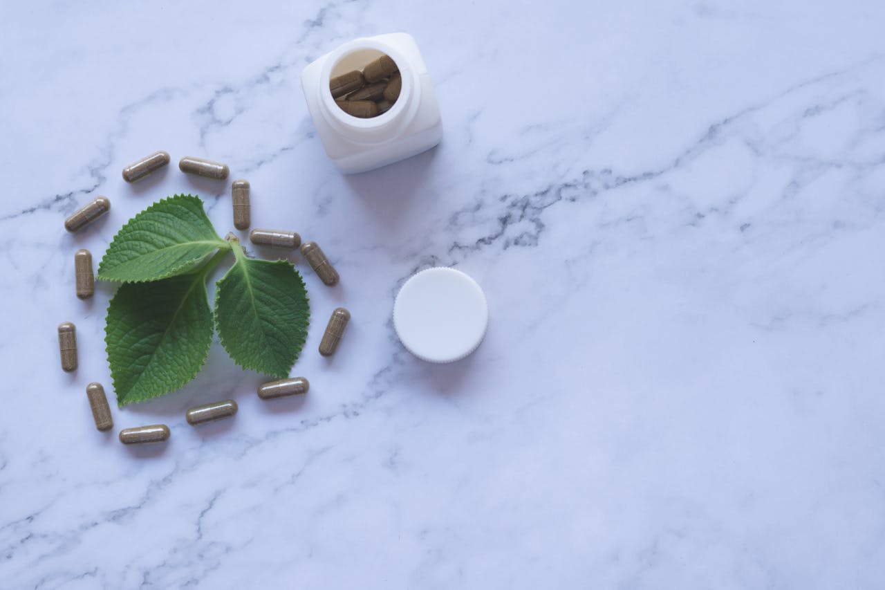 Flat lay of herbal capsules, leaves, and bottle on marble surface, symbolizing natural wellness.